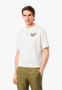 Camiseta de algodón blanca con mangas cortas, que presenta un logo negro de "Lacoste Golf" en el pecho. El modelo lleva puesta un pantalón de patrón verde oliva.