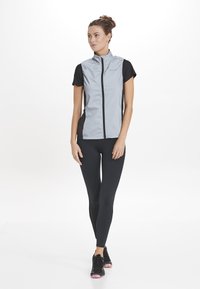 Grijze mouwloze jas met zwarte korte mouwen, ritssluiting aan de voorkant en zijpanelen; gecombineerd met zwarte leggings en zwarte sneakers met roze accenten.