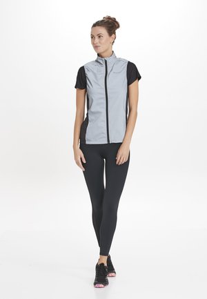 Graue ärmellose Jacke mit schwarzen kurzen Ärmeln, Frontreißverschluss und Seitenpaneelen; kombiniert mit schwarzen Leggings und schwarzen Sneakers mit pinken Akzenten.