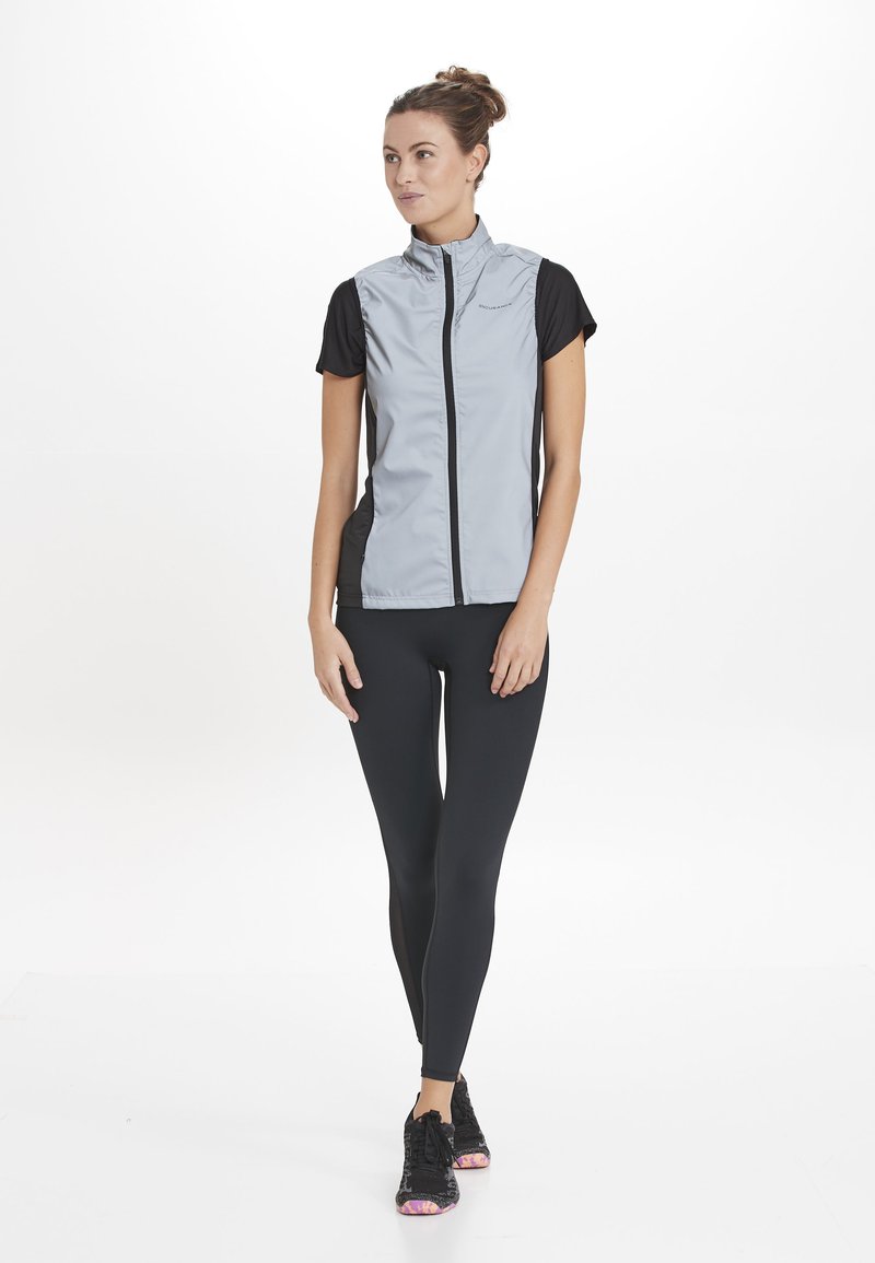 Grijze mouwloze jas met zwarte korte mouwen, ritssluiting aan de voorkant en zijpanelen; gecombineerd met zwarte leggings en zwarte sneakers met roze accenten.
