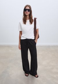 Witte T-shirt met korte mouwen, ontspannen zwarte wijde broek en donkere zonnebril. Bruine tas over de schouder. Gladde texturen, minimalistisch ontwerp.