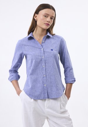 Light blue button-up shirt made of textured fabric, featuring a subtle embroidered bird logo and rolled sleeves, paired with white pants.