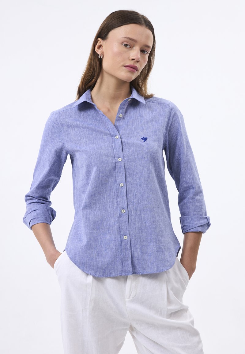 Camicia azzurro chiaro button-up realizzata in tessuto testurizzato, con un discreto logo di uccello ricamato e maniche arrotolate, abbinata a pantaloni bianchi.