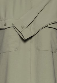 Manche de chemise vert clair repliée sur le corps de la chemise, montrant le poignet boutonné et les détails de couture.