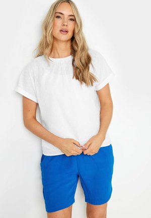 Młoda kobieta o długich blond włosach, ubrana w białą bluzkę z krótkim rękawem i jaskrawo niebieskie spodenki, na jednolitym białym tle.