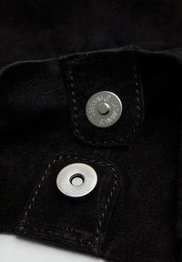 Tessuto in suede nero con due bottoni a pressione in metallo argentato e cuciture a contrasto. La vista ravvicinata mette in evidenza la texture e la finitura del materiale.