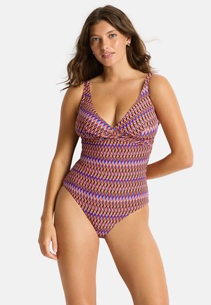 Kobieta z falującymi brązowymi włosami, ubrana w jednoczęściowy strój kąpielowy z kolorowym geometrycznym wzorem w odcieniach czerwieni, błękitu i bieli.