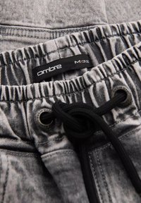 Cintura elástica de mezclilla gris con cordón negro atado en un nudo, mostrando una etiqueta negra etiquetada "ombre M | 32" dentro de la cintura.