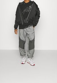 Personne portant une veste zippée noire sur un sweat à capuche graphique noir, un pantalon de jogging gris et noir, et des baskets blanches avec des accents rouges.