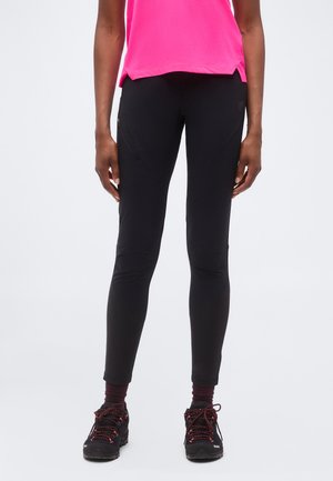 Osoba nosząca czarne dopasowane spodnie, ciemne buty trekkingowe z różowymi sznurówkami i jasnoróżowy krótki top na jednolitym tle.