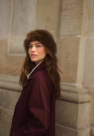 Femme aux longs cheveux bruns portant un chapeau en fourrure marron et un manteau bordeaux foncé, debout contre un mur de pierre beige, regardant la caméra.