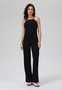 Schwarzer Jumpsuit mit schmalen Trägern, tailliertem Oberteil und weitem Hosenbein, aus einem glatten Stoff. Das Modell trägt sportliche Schuhe. Schlichtes Design.