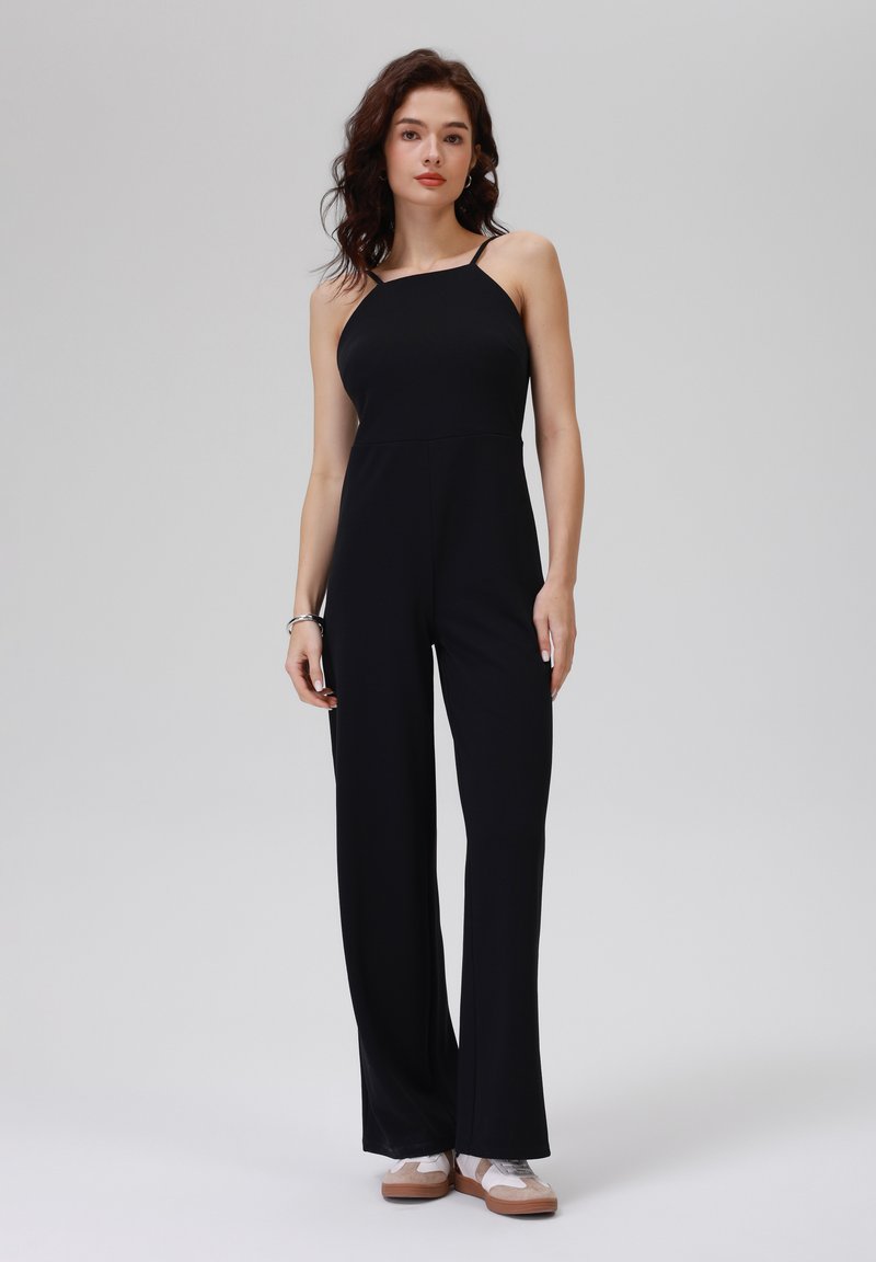 Schwarzer Jumpsuit mit schmalen Trägern, tailliertem Oberteil und weitem Hosenbein, aus einem glatten Stoff. Das Modell trägt sportliche Schuhe. Schlichtes Design.