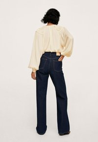 Blusa crema con maniche a palloncino, abbinata a jeans scuri a vita alta. La blusa presenta dettagli testurizzati lungo le spalle e la schiena.