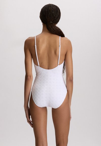 Γυναίκα με πλεξούδες, φορούσα λευκό ολόσωμο μαγιό με λεπτές ρυθμιζόμενες τιράντες, φαίνεται από πίσω σε απλό φόντο.