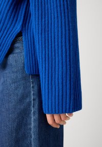 Bras droit portant une manche de pull côtelé bleu à côté d'une main, associé à un jean en denim bleu.