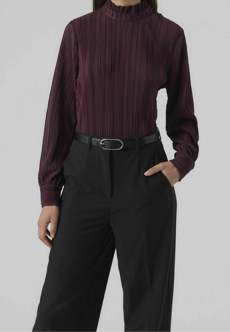 Femme portant un chemisier prune à manches longues et col montant avec des plis verticaux, un pantalon noir à jambes larges et une ceinture noire avec une boucle argentée.