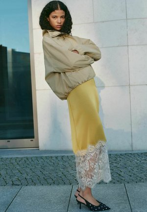 Woman with curly hair wearing beige bomber jacket, yellow skirt with white lace hem, and black polka-dot heels, standing outdoors on stone pavement.