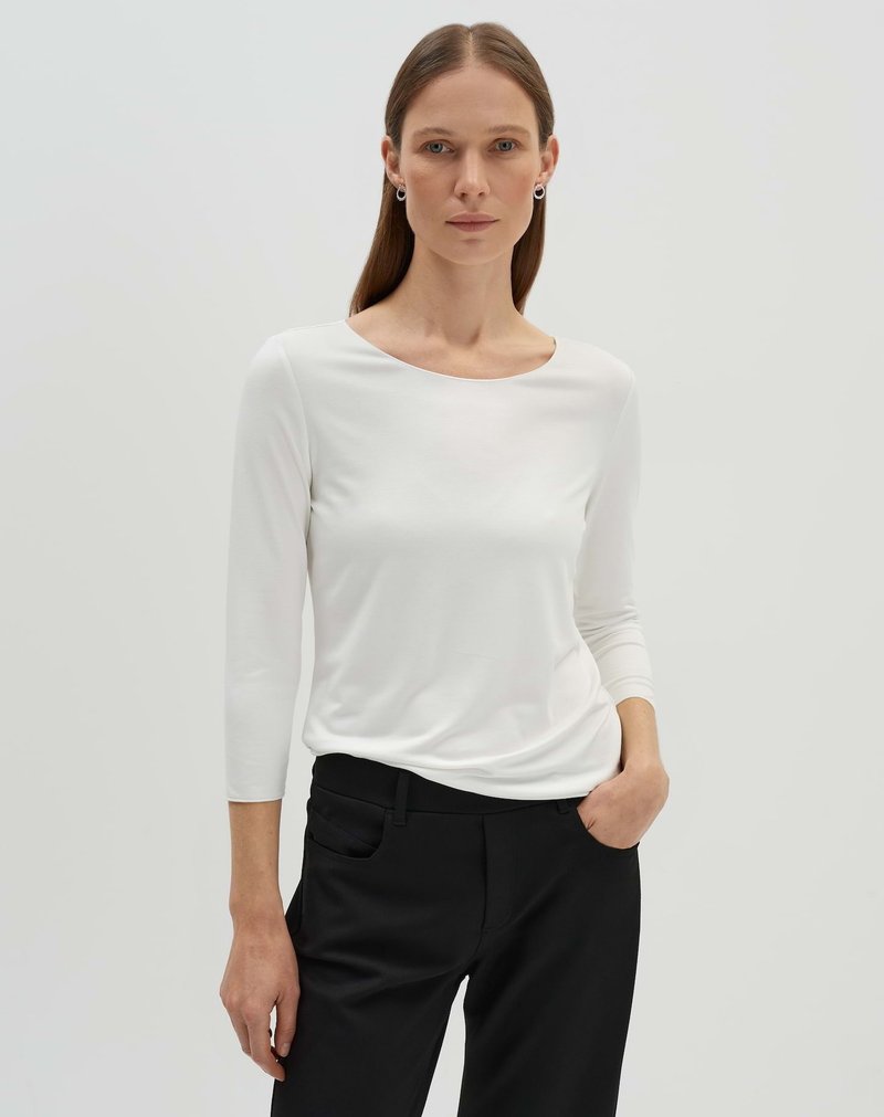 Haut blanc à manches longues avec un col bateau, en tissu lisse, associé à un pantalon noir ajusté avec poches latérales.