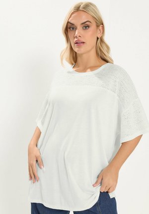 Blonde vrouw met een los wit shirt met gestructureerde gebreide mouwen en een blauwe spijkerbroek, staand tegen een effen lichte achtergrond.