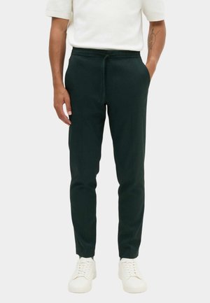 Mand iført sorte slim-fit joggingbukser med snørebånd i taljen, hvide sneakers og en hvid kortærmet skjorte, stående mod en ensfarvet baggrund.