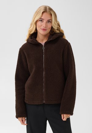 Woman with blonde hair wearing a dark brown zip-up fleece jacket with a hood and black pants against a plain light background.