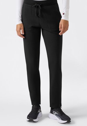 Osoba ubrana w czarne joggery slim-fit z przednimi kieszeniami, czarno-białe buty sportowe oraz białą koszulę z długim rękawem i logo na nadgarstku.