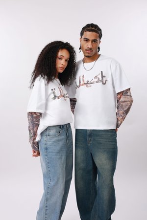 Dwie osoby stoją obok siebie, ubrane w białe koszulki z nadrukiem na długich, kamuflażowych rękawach oraz luźne niebieskie jeansy, na jednolitym tle.