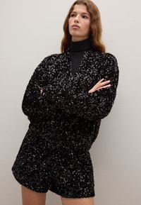 Black sequined bomber jacket with a zip front and matching shorts, featuring a textured surface and relaxed fit, worn over a turtleneck top.