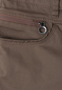 Gros plan d'une poche de pantalon en tissu marron avec une fermeture éclair horizontale et un tire-zip rond en métal.