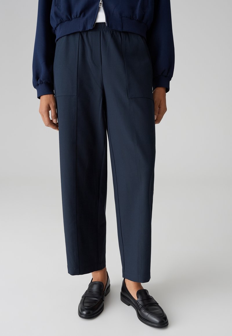 Personne portant un pantalon bleu marine à jambes droites avec des poches avant, une veste bleu marine et des mocassins en cuir noir, debout sur un fond uni.