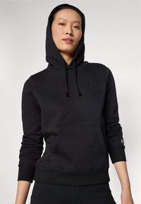 Svart hoodie av mjukt material, med en framficka, dragsko i huvan och logotypdetalj på bröstet och ärmen. Fitted muddar.