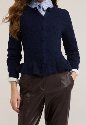 Femme portant un cardigan bleu marine boutonné avec un bas à volant, une chemise rayée à col et un pantalon en cuir marron foncé, les mains dans les poches.