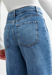 Pantalones vaqueros azules de mezclilla con una pretina alta, que cuentan con bolsillos traseros curvados y una textura ligeramente desgastada. Detalles de costuras recorren los lados.