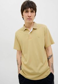 Šviesiai rudos spalvos polo marškiniai su trumpomis rankovėmis, klasikinis apykaklė ir subtilus logotipas krūtinės srityje. Lygi tekstūra, laisvas fitas ir minimalus dizainas.