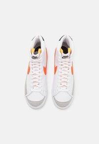 Höga sneakers med ett vitt läderöverdrag, orange Swoosh, rosa snören, grå mockadetaljer och svart branding på tungan.