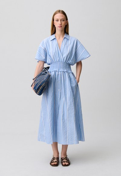 Donna che indossa un vestito midi a righe azzurre con vita arricciata, tiene una borsa intrecciata blu e indossa sandali scuri con cinturini, in piedi davanti a uno sfondo semplice.