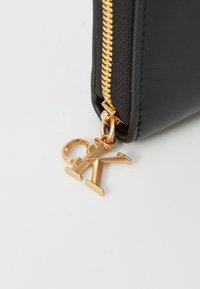 Coin du sac en cuir noir avec une fermeture éclair en or et un charme logo en or affichant "CK" attaché au tire-zipp.