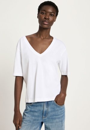 Junge Person trägt ein lockeres weißes V-Ausschnitt-T-Shirt mit kurzen Ärmeln und hellblaue Jeans, eine Hand in der Tasche, vor einfachem Hintergrund.