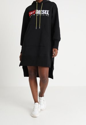 Persoon draagt een zwarte Diesel hoodie-jurk met gele trekkoorden en witte sneakers, staand tegen een effen witte achtergrond.