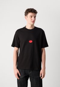 Black cotton T-shirt featuring a round neckline, short sleeves, and a red circular logo with "HUGO" at the center. Worn with dark jeans.