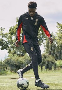 Puma idrottsträningssuit i svart med röda accenter, som har en logotyp, en jacka med dragkedja och smalnar byxor, kombinerat med svarta fotbollsskor och en fotboll.