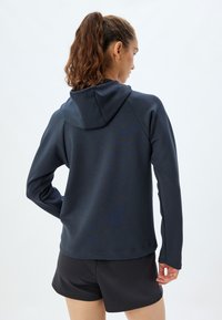 Sudadera deportiva azul oscuro con capucha, mangas largas, textura suave y un corte relajado, combinada con pantalones cortos negros.