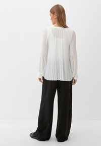 Haut plissé blanc à manches longues, col rond et poignets boutonnés ; associé à un pantalon noir à jambes larges et des baskets noires.