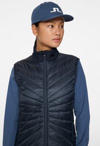 Siniharmaa eristetty liivi, jossa on viistokuviointi, korkea kaulus ja koko edestä avattava vetoketju. Yhdistetty yhteensopivan logolla varustetun pipon kanssa.