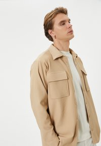 Chemise boutonnée beige clair en tissu texturé, avec deux poches sur la poitrine et un design à col, portée avec un tee-shirt blanc à col rond en dessous.