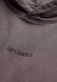 Sudadera con capucha gris hecha de algodón texturizado, con un logo "SUPERDRY" ligeramente elevado en hilo a juego a lo largo del pecho.