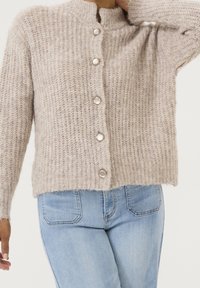 Cárdigan de punto beige claro con cuello alto, que presenta cinco botones grandes y redondos y un dobladillo ligeramente corto. Tejido suave y texturizado.