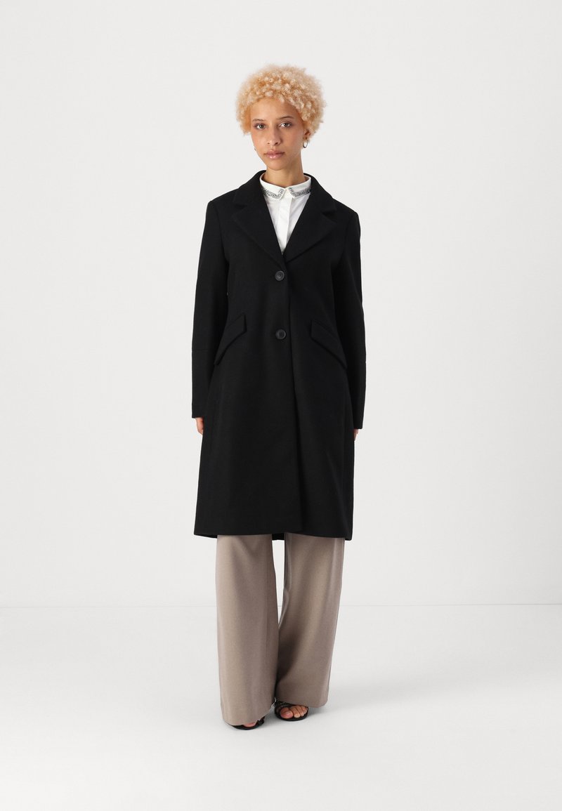Cappotto di lana nero con revers a scialle, due bottoni frontali e tasche laterali. Design semplice, lunghezza media, indossato sopra una camicia a colletto chiara e pantaloni.