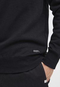 Svart sweatshirt med ribbat avslut och ärmslut, tillverkad av mjukt material. Har en liten svart etikett med vit text nära nederkanten till vänster.